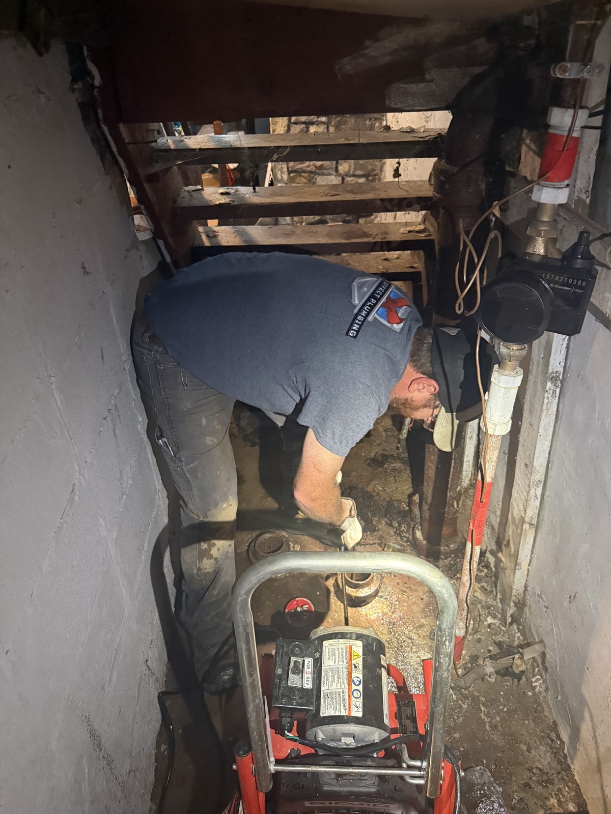 Lake Effect Plumbing at work, in a company shirt, running a RIDGID drain machine down a floor drain in a tight old basement