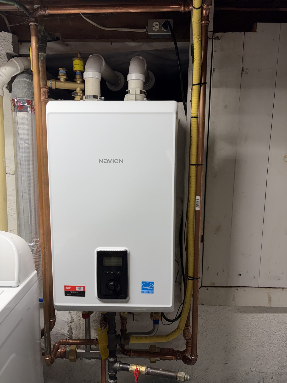 A Navien tankless water heater installed on a basement wall with new copper water lines, a yellow gas line and a Lake Effect Plumbing sticker on the unit