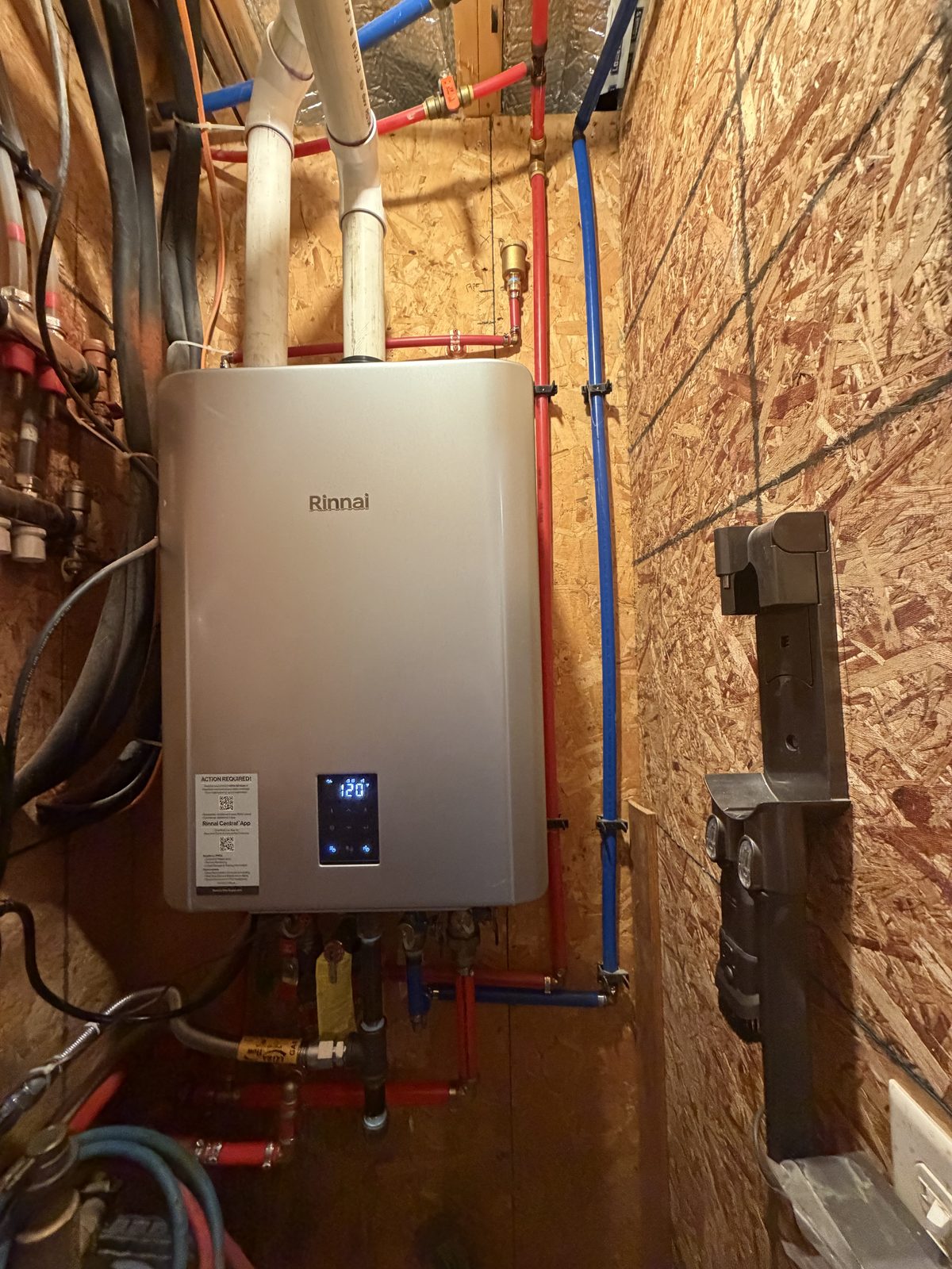 A Rinnai tankless water heater installed in a utility closet with red and blue PEX water lines and white PVC venting